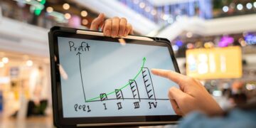 A manager using tablet to see business growth prediction graph of 2021 on screen with blur background of department store interior. Business successful concept. Selective focus at human hand part.  Our Services trades 360x180