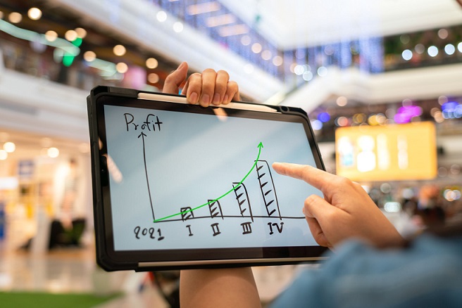 A manager using tablet to see business growth prediction graph of 2021 on screen with blur background of department store interior. Business successful concept. Selective focus at human hand part.  Capital Raising and Investment Management trades